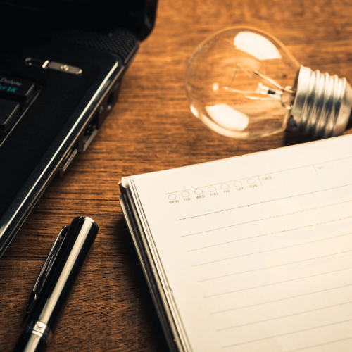Next to a laptop, there is notebook with a lightbulb